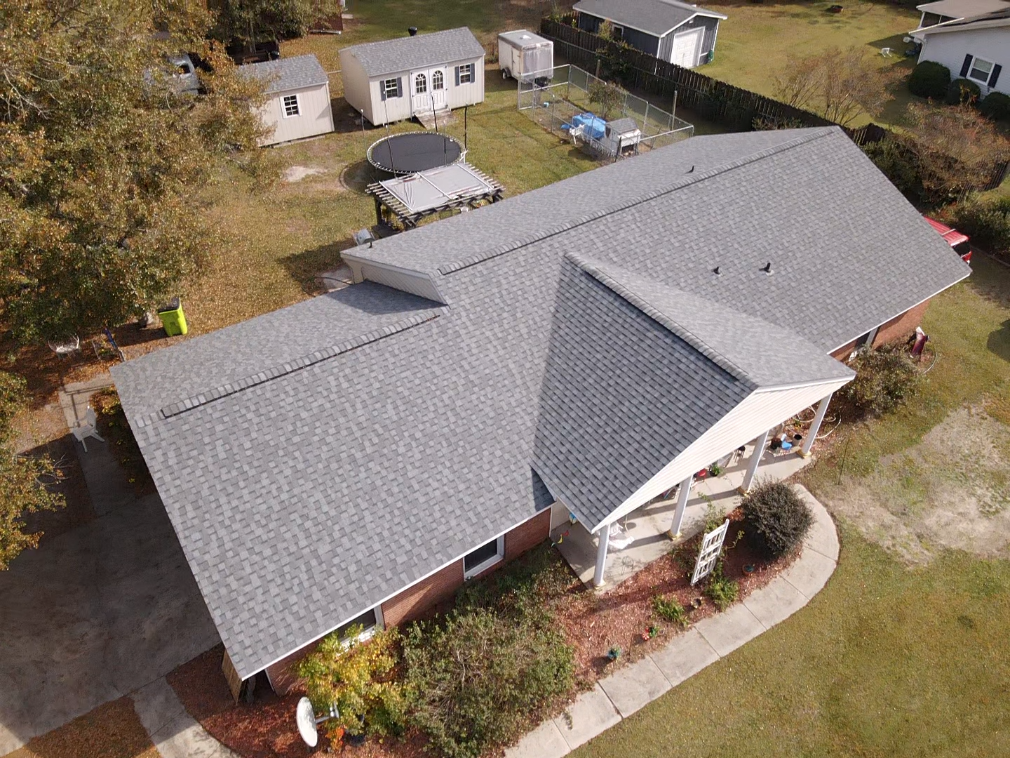 Aerial view of a gray-shingled roof on a brick house with a small yard, trees, and small buildings.