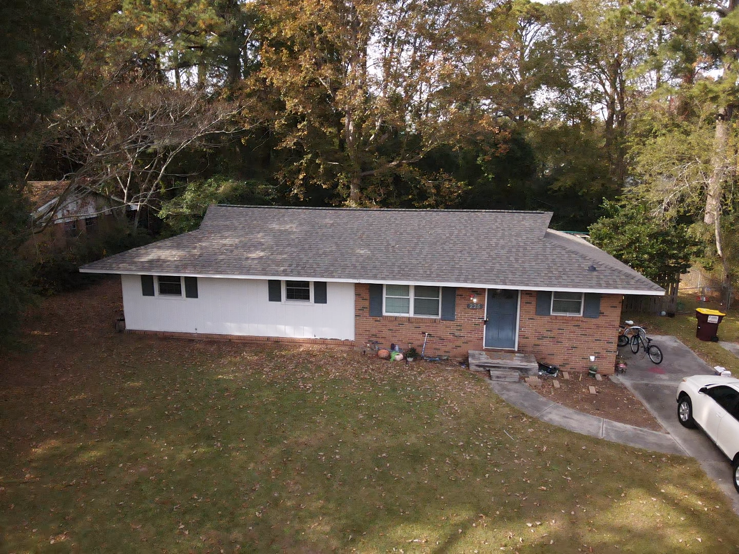 Ranch-style house with brick and white siding. Trees and grass surround it. Car and bicycle are in the driveway.