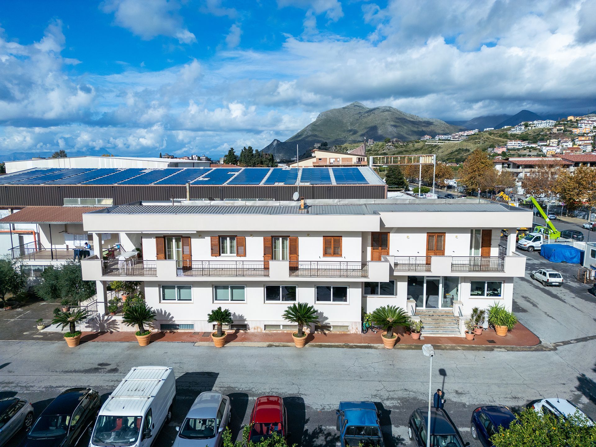 Edificio bianco a due piani con balconi, pannelli solari sul tetto, auto parcheggiate di fronte, montagne sullo sfondo, cielo azzurro.