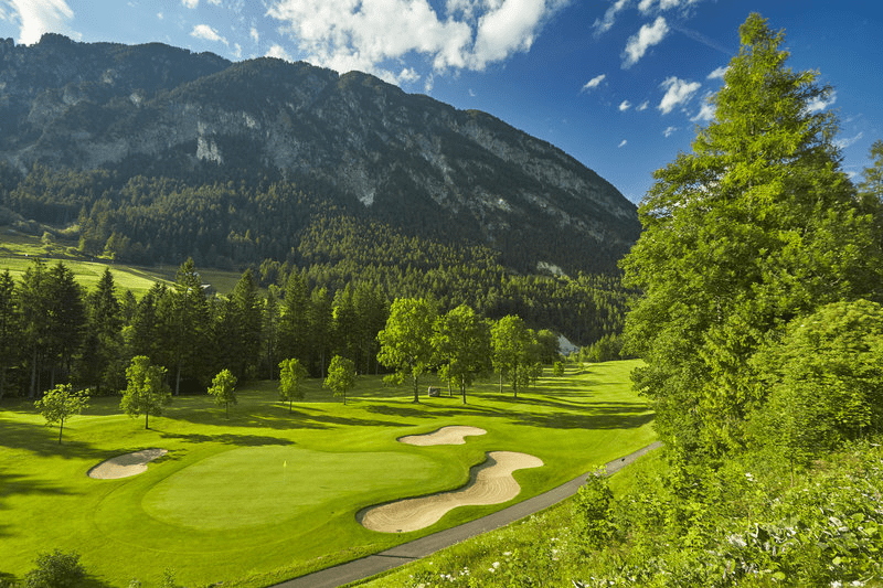 Ein Golfplatz mit Bergen im Hintergrund und Bäumen im Vordergrund.