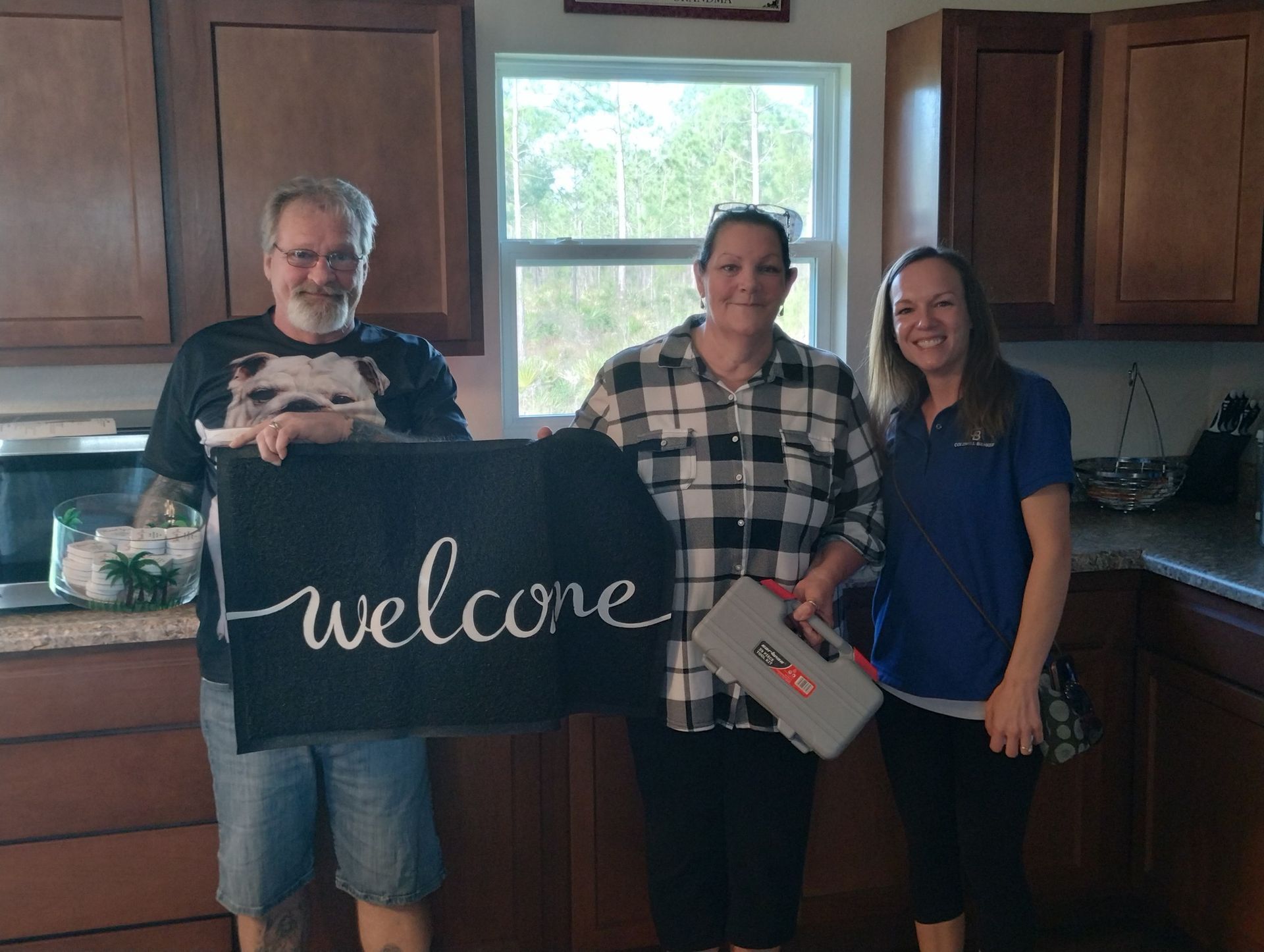 Three people holding a welcome sign and tool box in a kitchen, smiling.