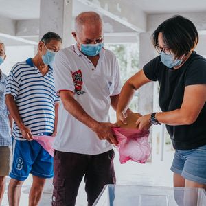 People in masks receiving supplies in a light-filled building. A woman hands a bag to a man.