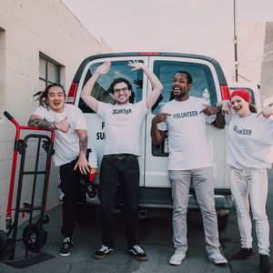 Four volunteers in white shirts in front of a van; one with arms raised, another with a dolly.