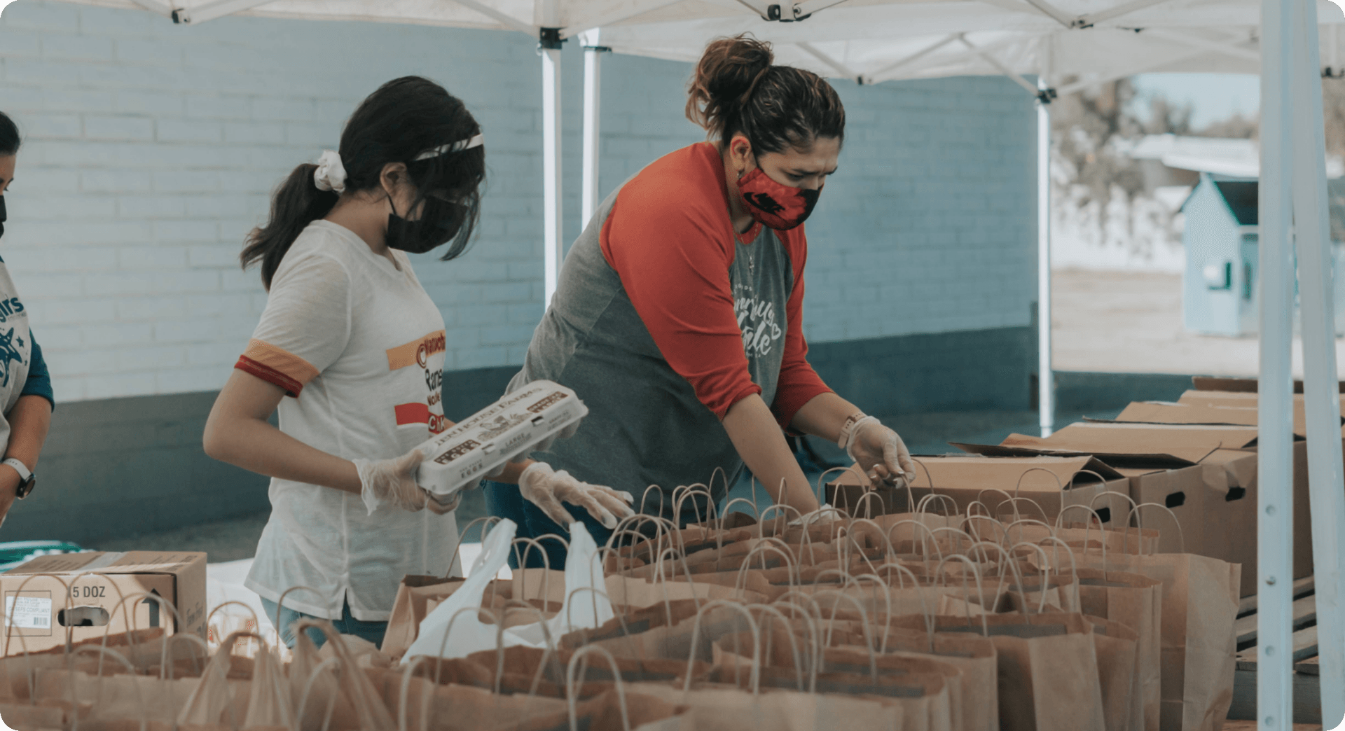 Two masked women pack groceries into paper bags at an outdoor market.
