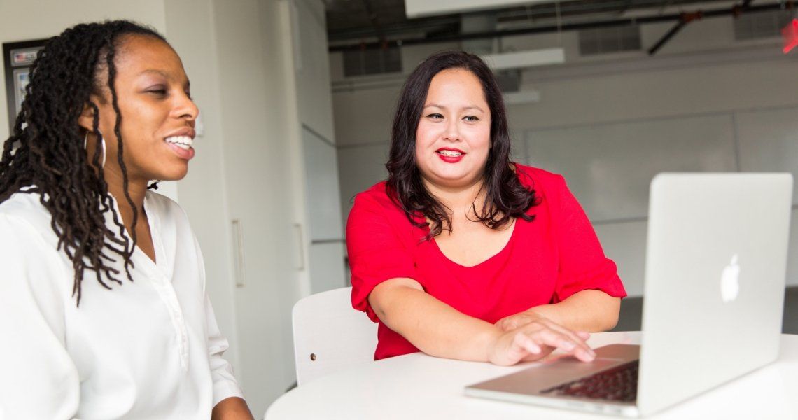 Two women at a table with a laptop. One in red top, smiling and typing. Other in white, smiling.