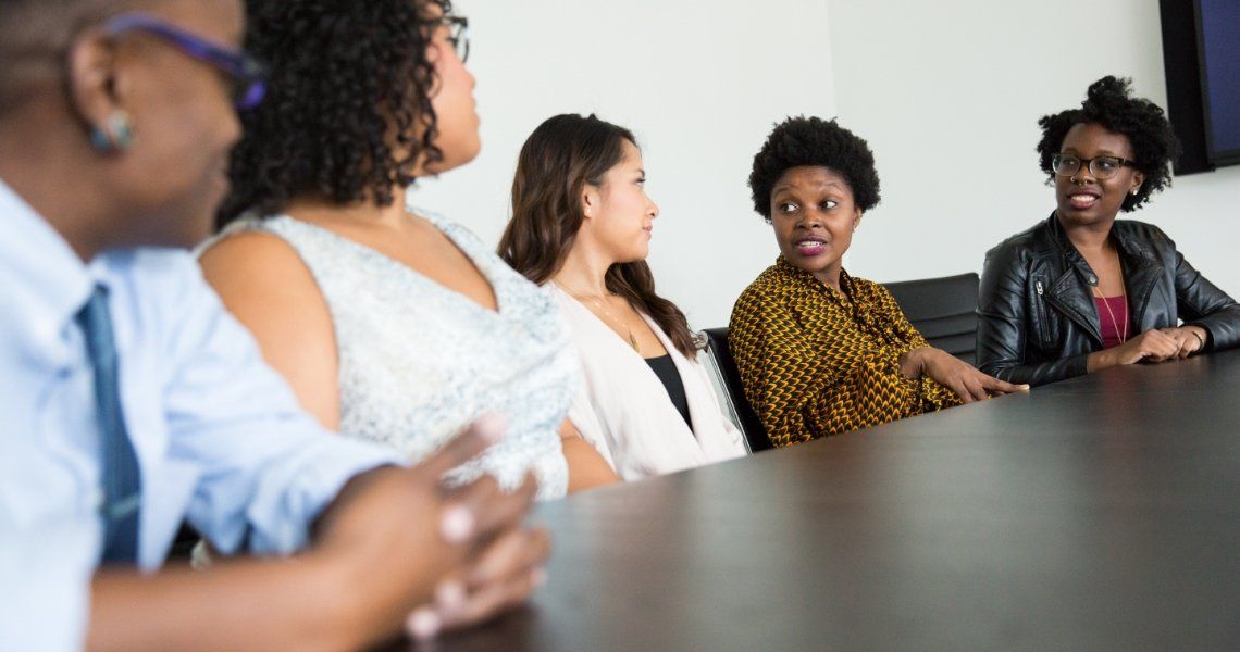 Group of five diverse women seated at a conference table, engaged in discussion.