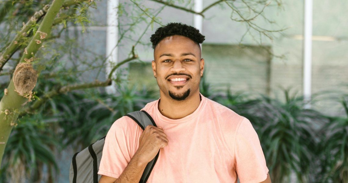 Smiling young man in a pink shirt, holding a backpack, standing outside near greenery.