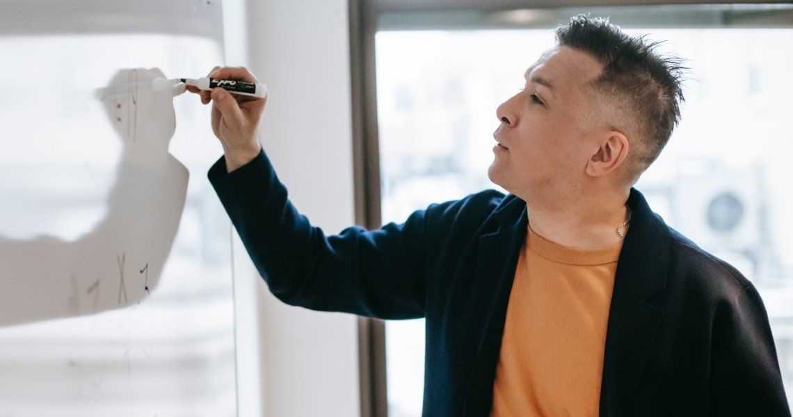 Man writing on a whiteboard, wearing an orange shirt and dark jacket, by a window.