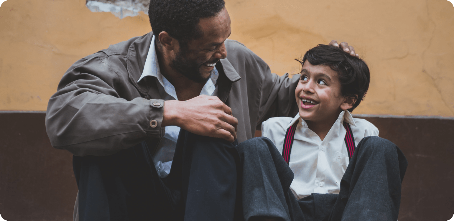 A smiling Black man with his arm around a smiling young boy, outside, against a yellow wall.
