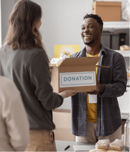 Person giving a donation box to a smiling man; indoor setting, likely a charity drive.
