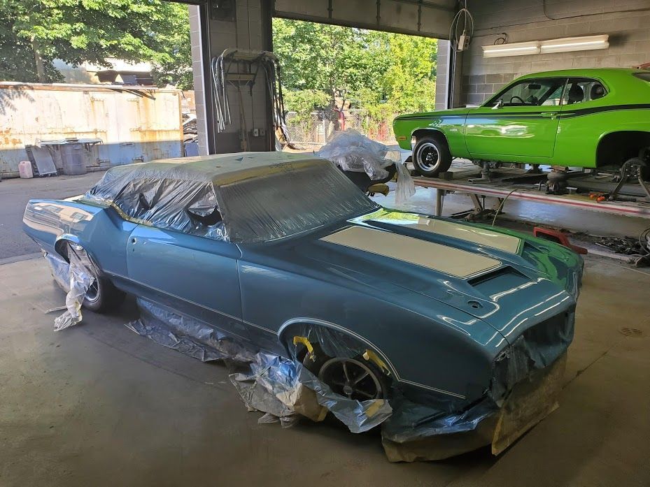 A blue car is being painted in a garage next to a green car.