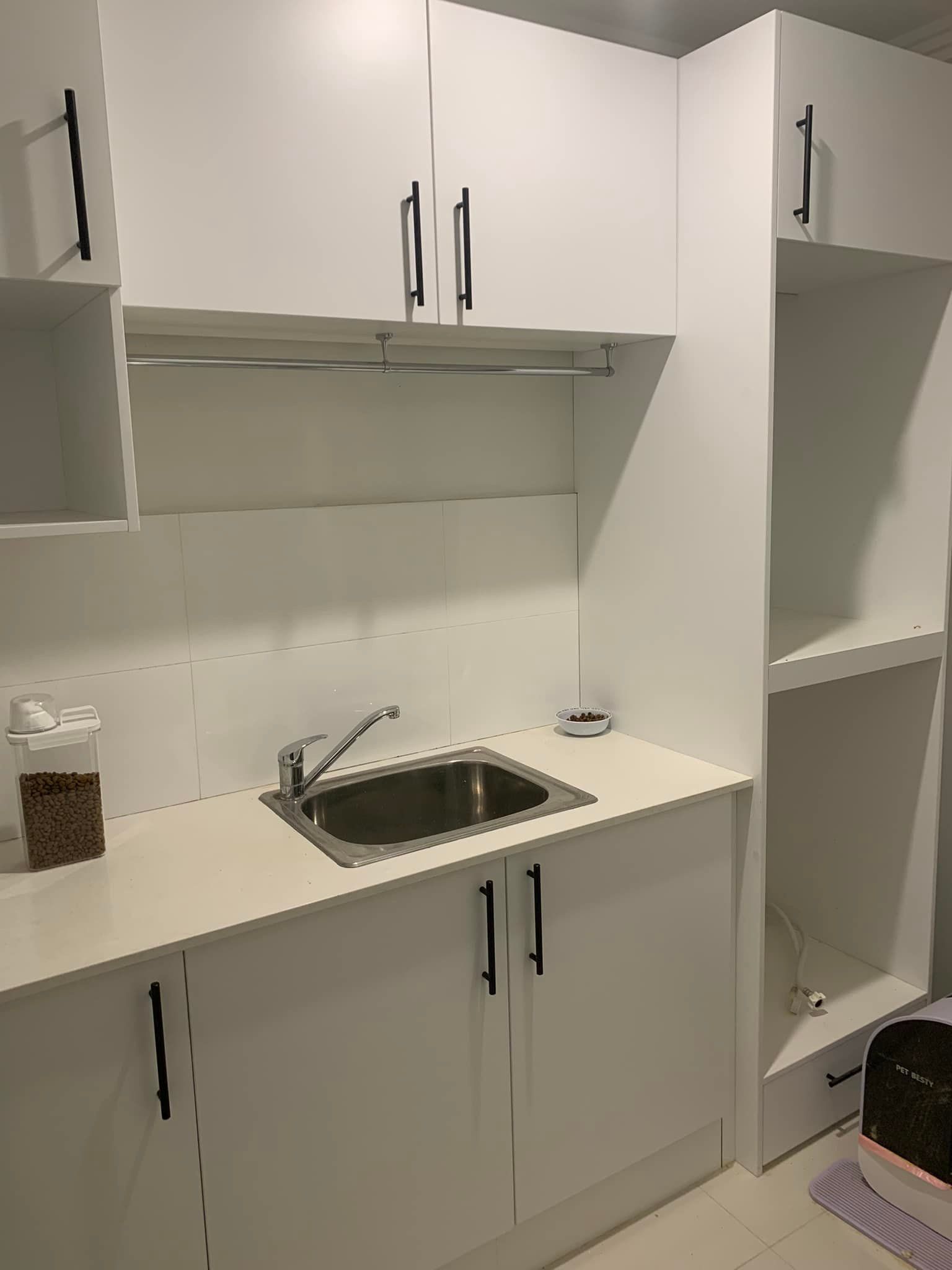 A kitchenette with white cabinets, a stainless steel sink, and a tall shelf unit with black handles on a white wall.