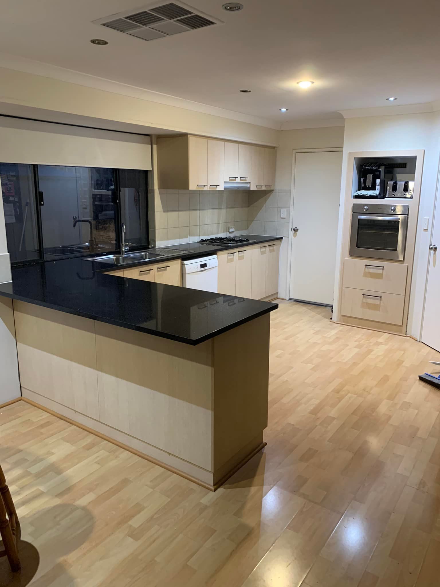 A modern kitchen with light-wood cabinets, black granite countertops, a built-in oven, and light-colored wooden floors.