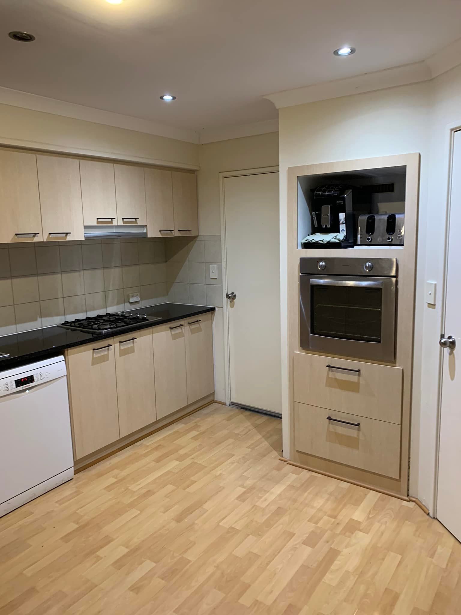 A kitchen with light wood cabinets, black countertops, a dishwasher, a built-in oven, and light wood flooring.