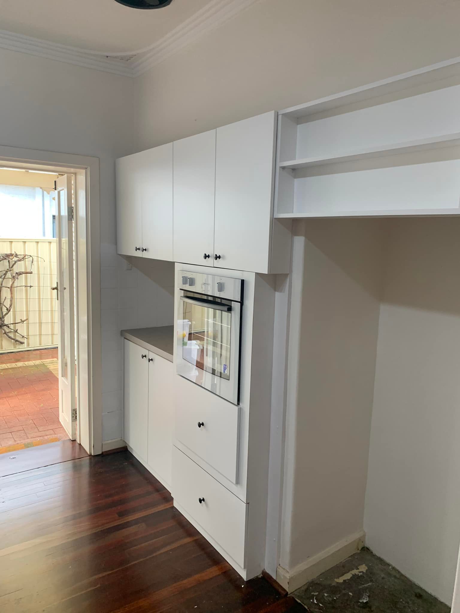 White kitchen cabinets with a built-in oven and open shelving next to a doorway with dark wooden floors.