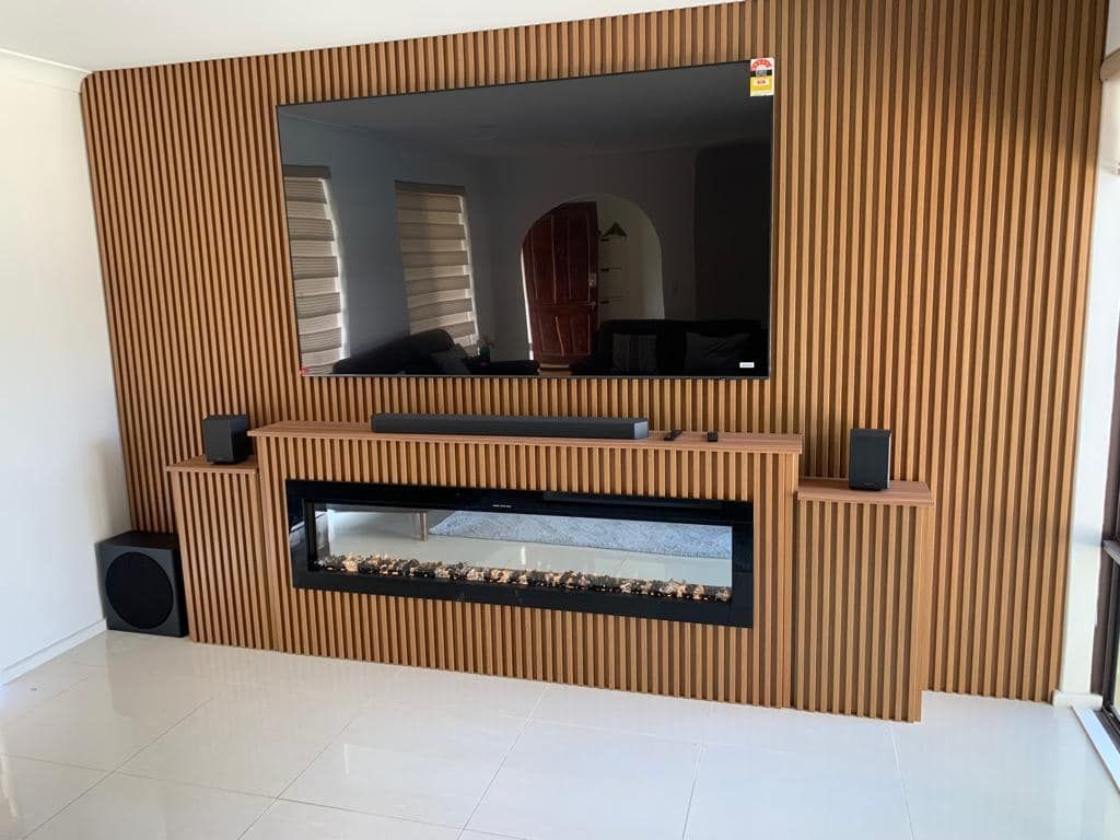 A wall-mounted TV sits above a wide fireplace, surrounded by vertical wood-slat paneling with speakers on a shelf below.