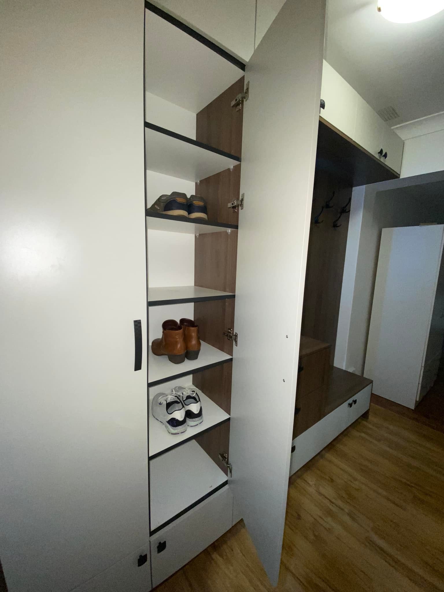 An open white cabinet with wood-toned interior shelves, storing pairs of shoes, located in an entryway with wood floors.