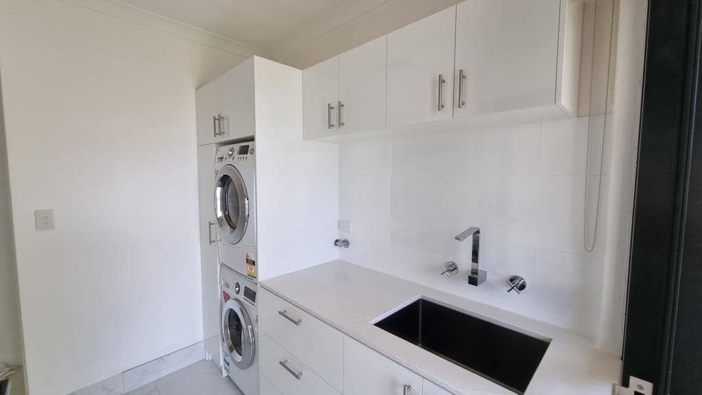 A modern laundry room with white cabinetry, a stacked washer and dryer, and a black sink basin on a white counter.