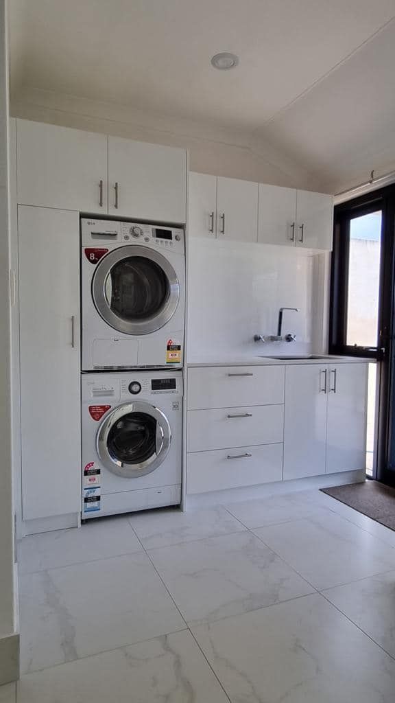 A laundry room with white cabinets, a stacked washer and dryer, a sink, and white tiled floors.