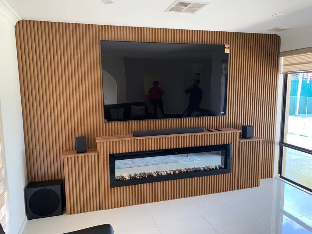 A wall-mounted TV above a linear fireplace, framed by wood slat paneling with speakers on flanking shelves.