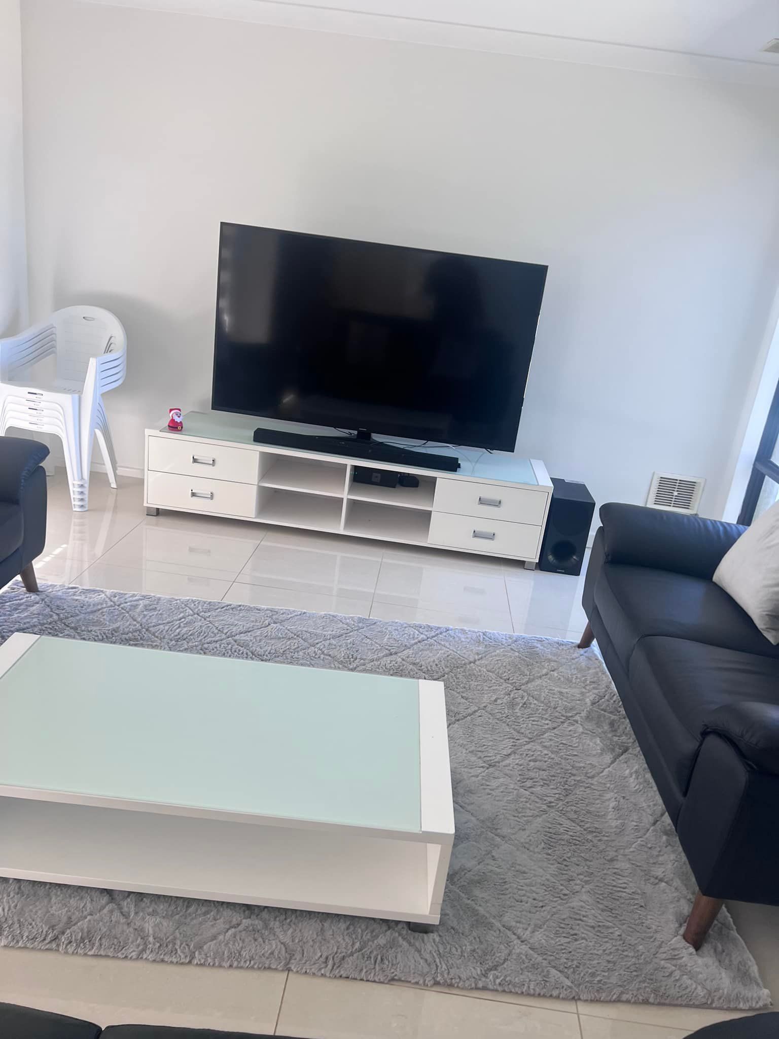 A living room with a large TV on a white console, a gray rug, a glass-top coffee table, and a dark sofa.