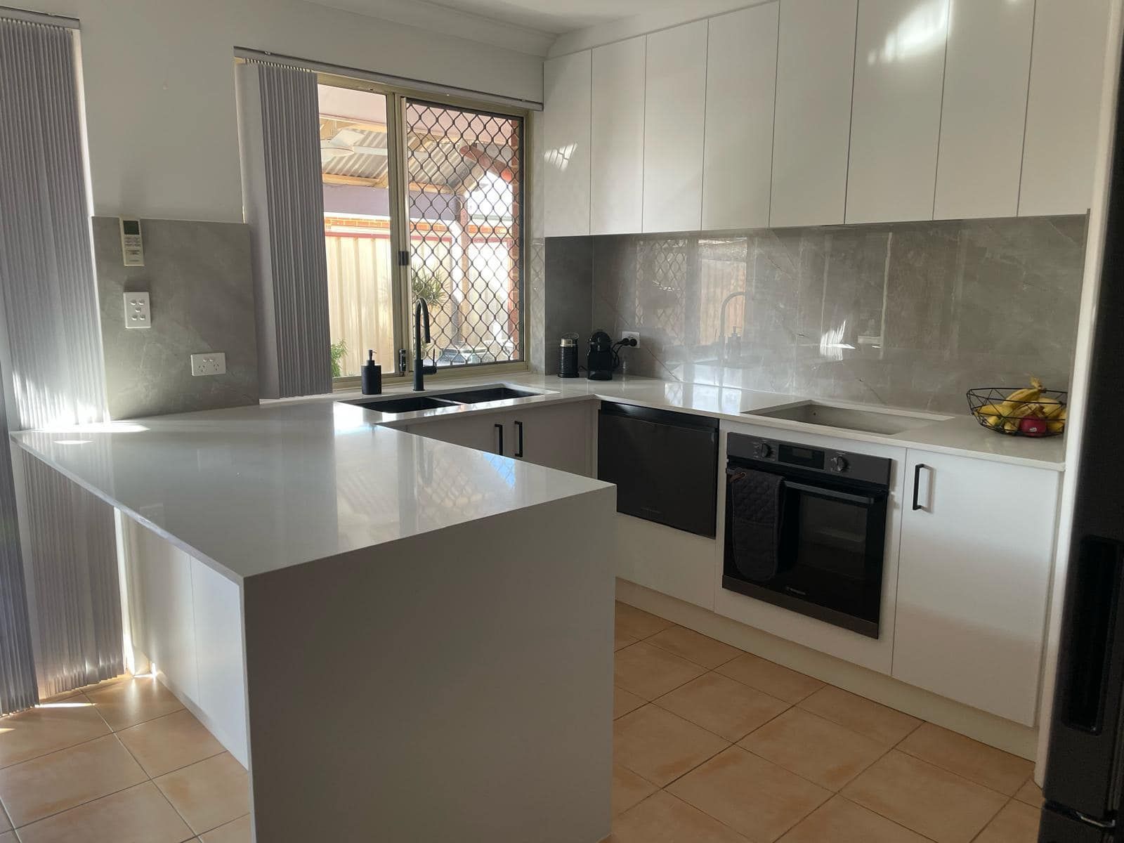 A modern kitchen featuring white cabinets, a light-colored island bench, grey tiled backsplash, and a window with a grill.