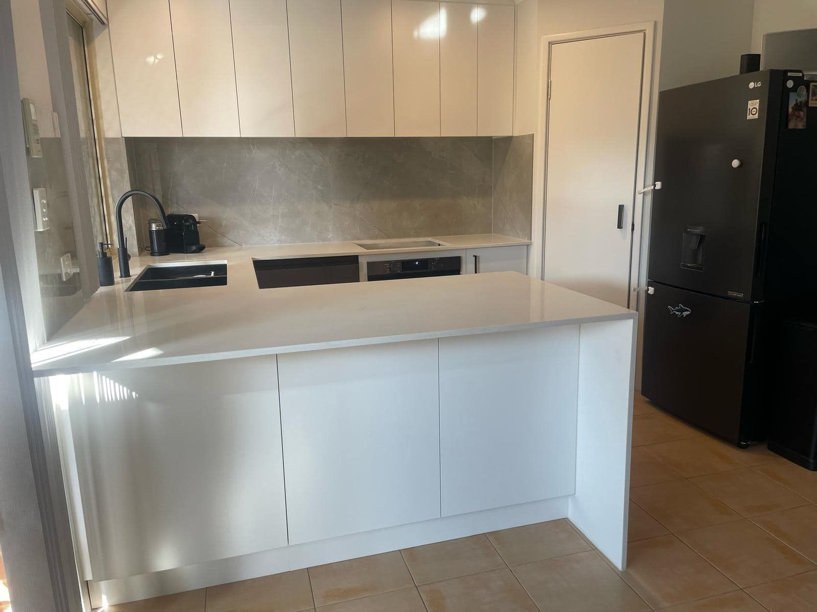 A modern kitchen with white cabinets, a large white breakfast bar, a concrete-look backsplash, and a tall black fridge.