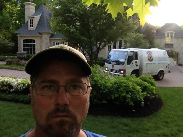 Man in cap selfie with house, white van in the driveway, and landscaping.