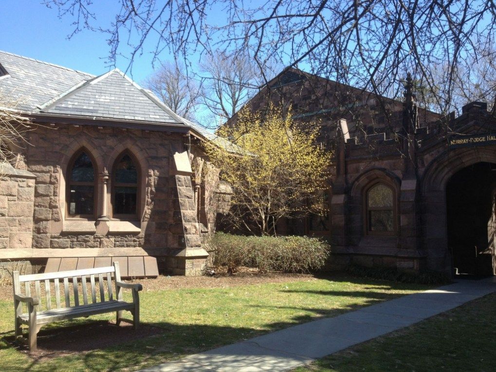 Stone buildings with a bench, green lawn, and flowering bush under a blue sky.
