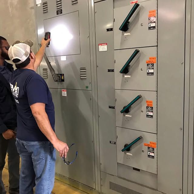 A technician in a navy shirt and white hat points a flashlight at a large gray industrial electrical panel.