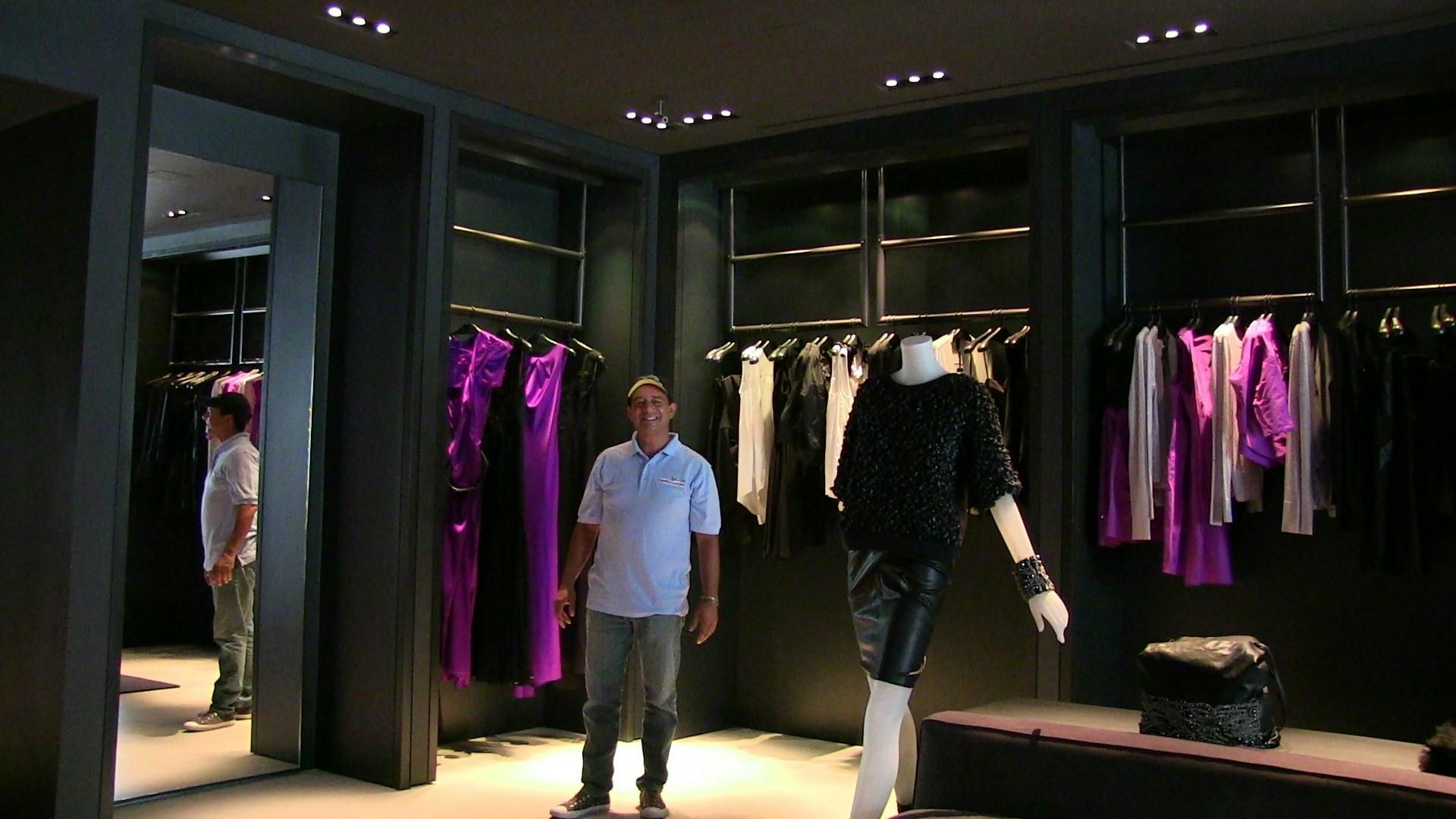 A man stands in a clothing store with dark shelving, displaying purple and white garments and a mannequin.