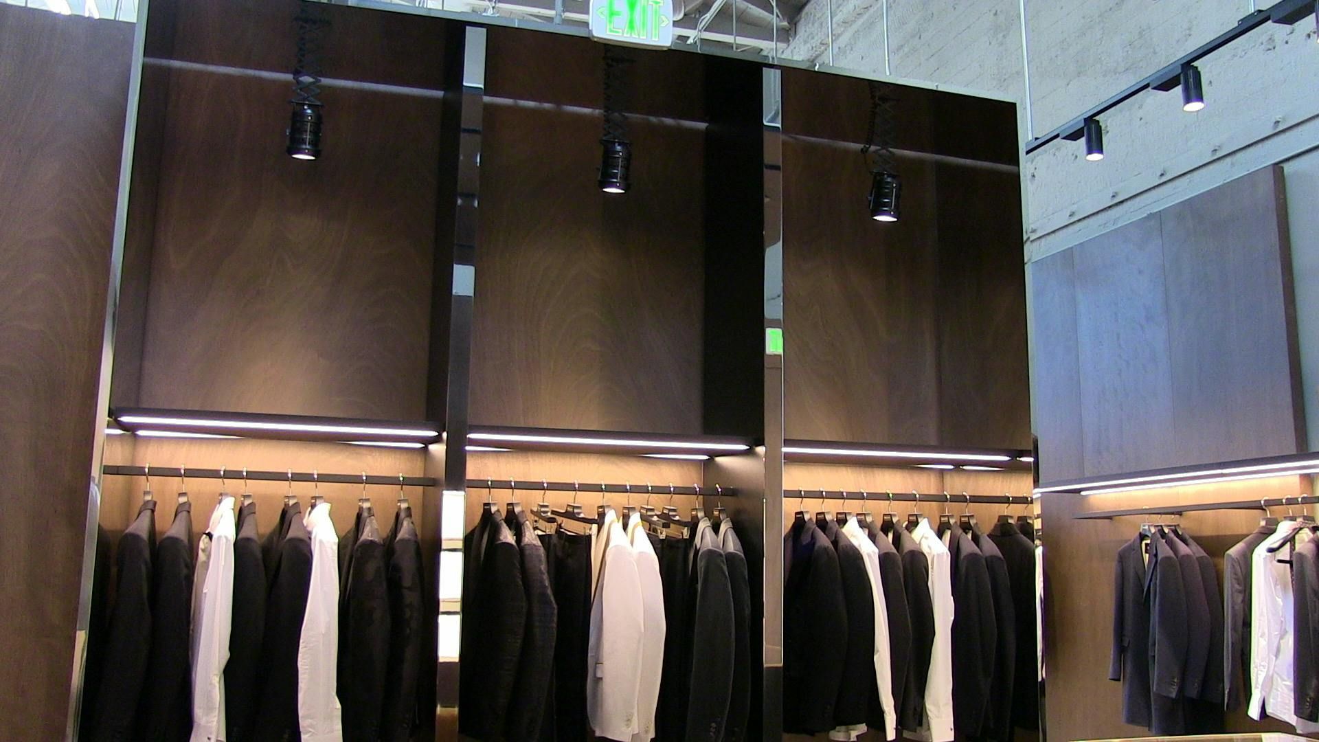 Rows of suits and shirts hang on racks under dark wooden display cabinets in a brightly lit clothing store.