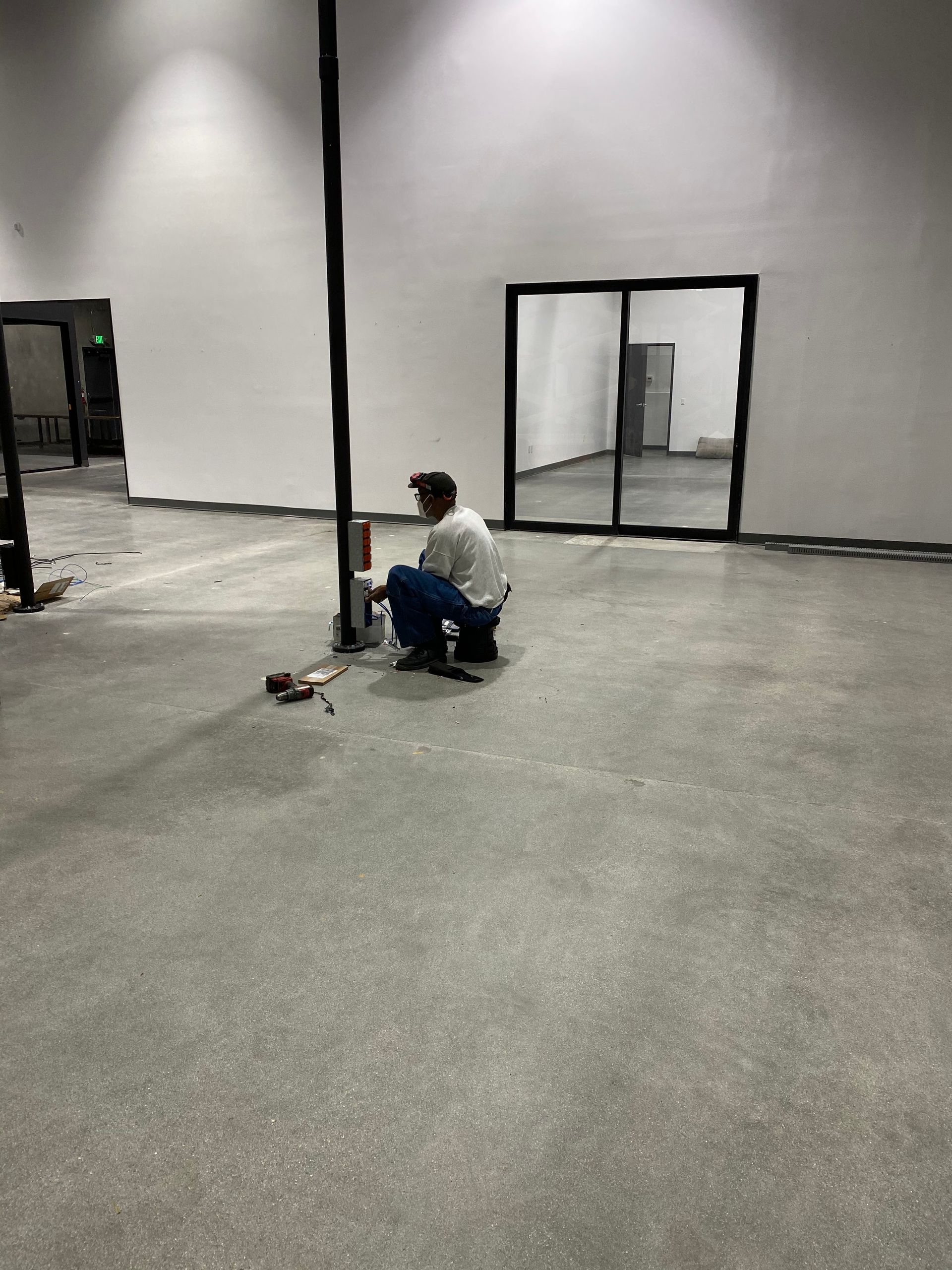 Person crouches beside a black pole in a large empty indoor space with gray carpet and white walls.