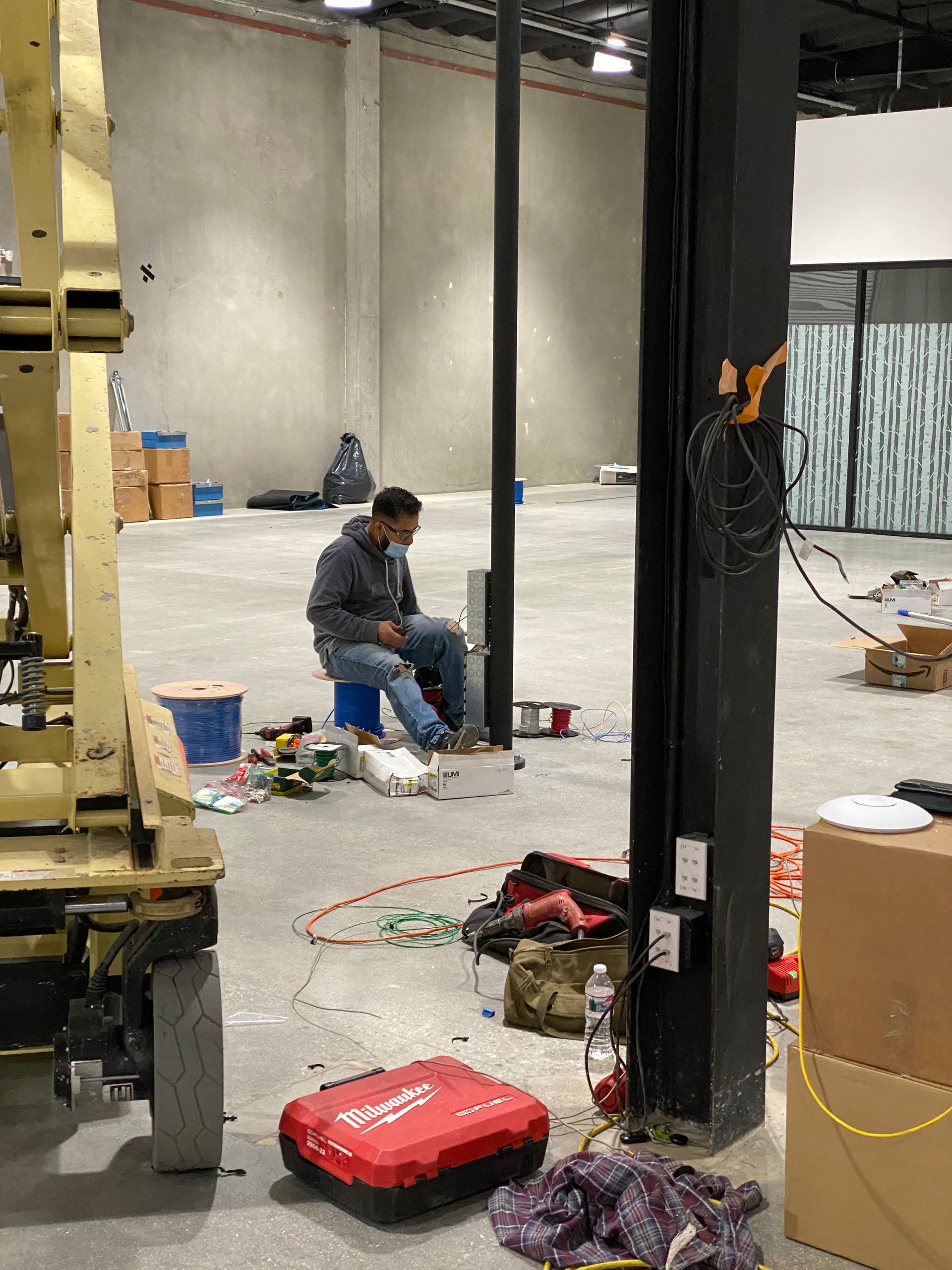 Worker kneeling on a concrete floor in a warehouse under construction, with tools and materials scattered nearby.