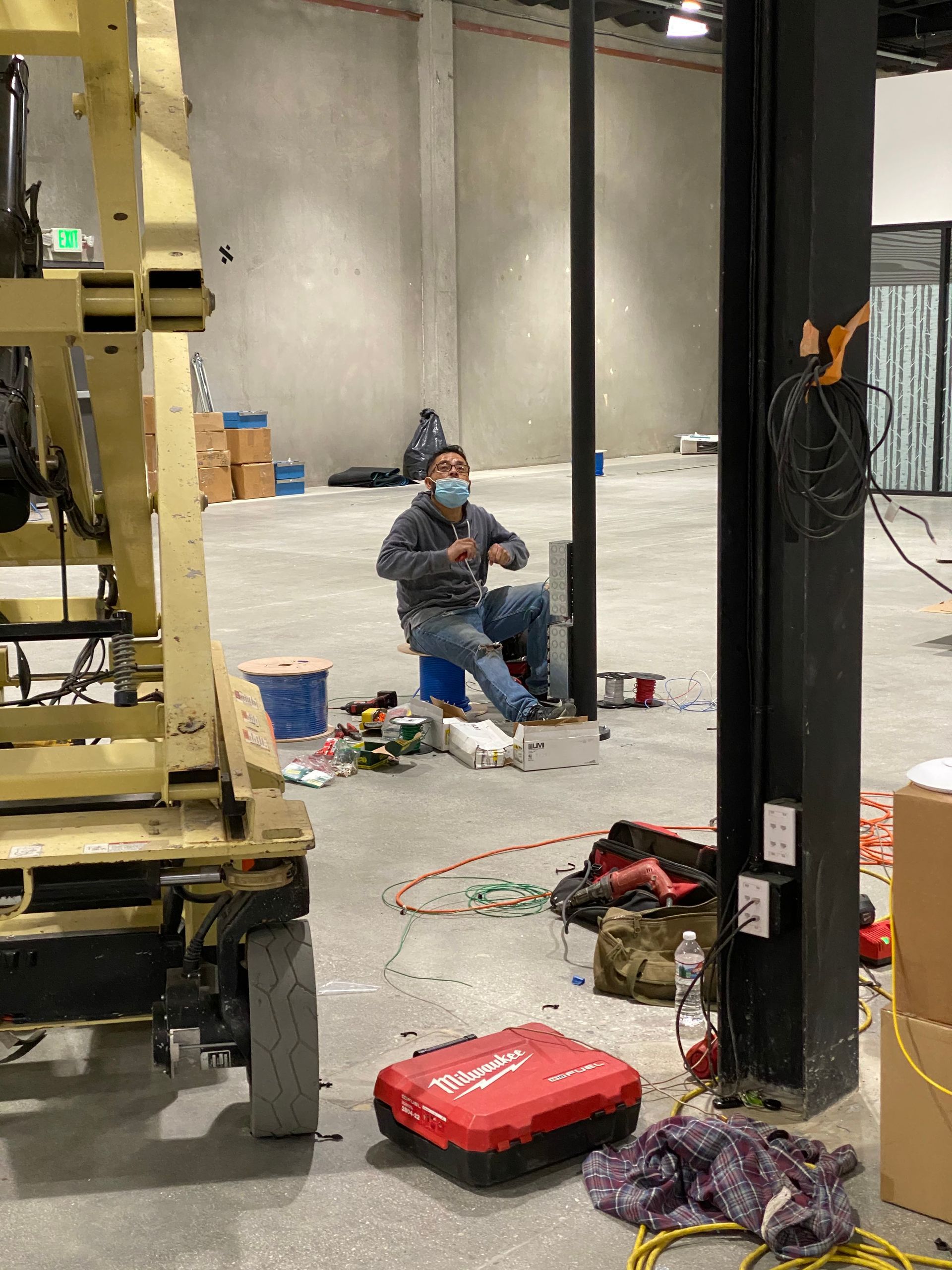 Worker seated in a warehouse beside a lift, tools and cables scattered on the floor.