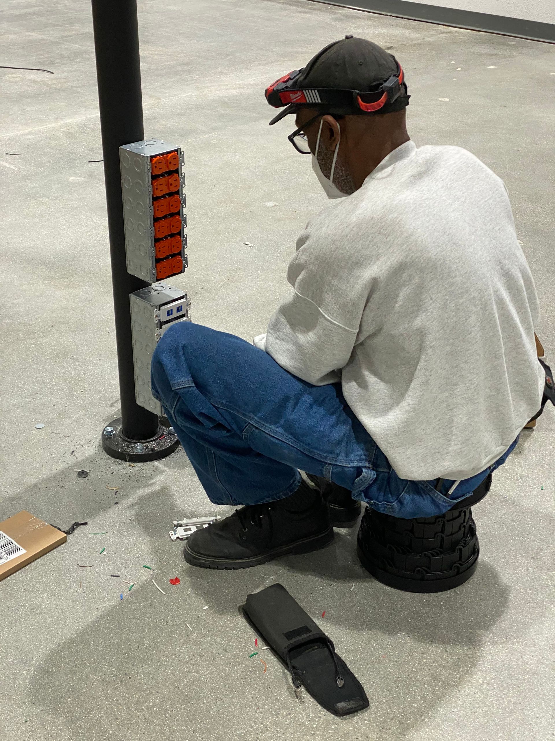 Man crouching beside a utility pole, adjusting a small orange device on a concrete floor.
