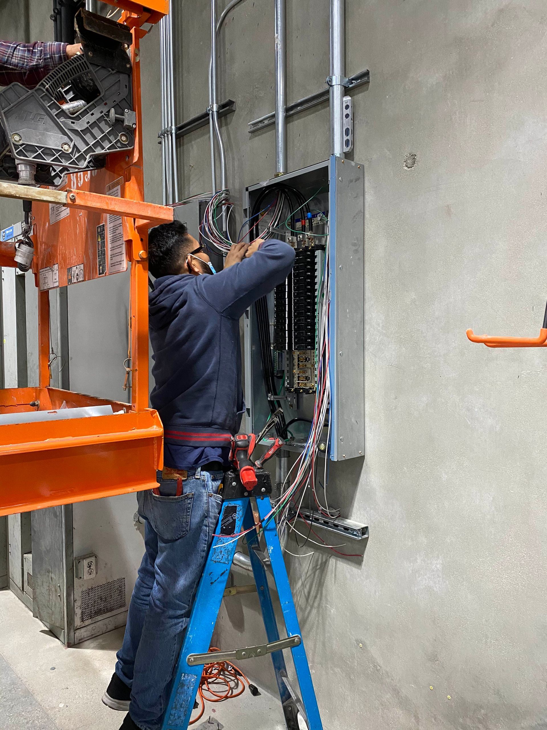 Worker in a blue jacket installing electrical wiring in a wall panel beside an orange lift.