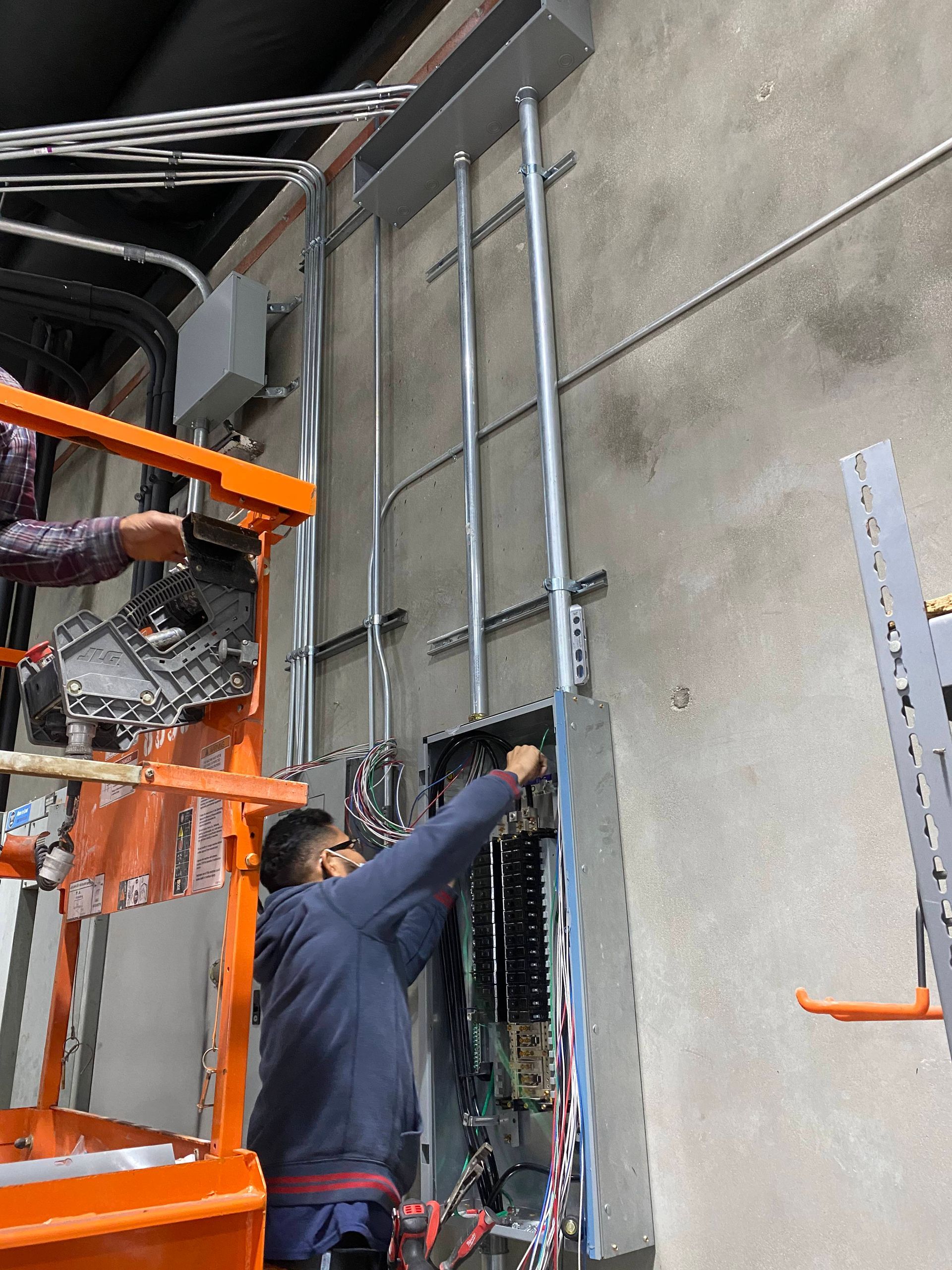 Worker in blue jacket pointing at a tall gray wall beside orange scaffolding in an industrial space