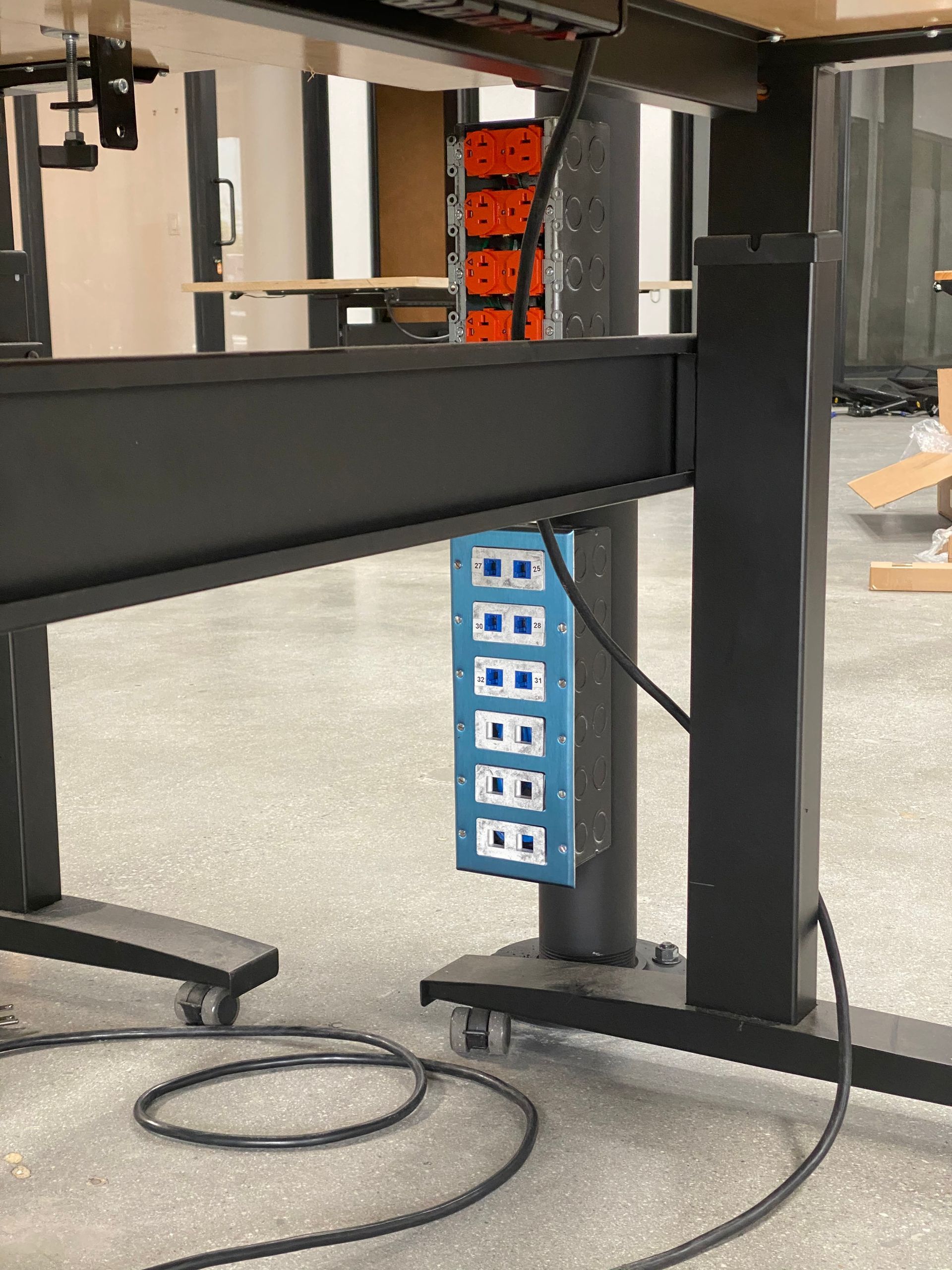 Blue power strip hanging under a black standing desk in a workshop setting, with cords on the floor