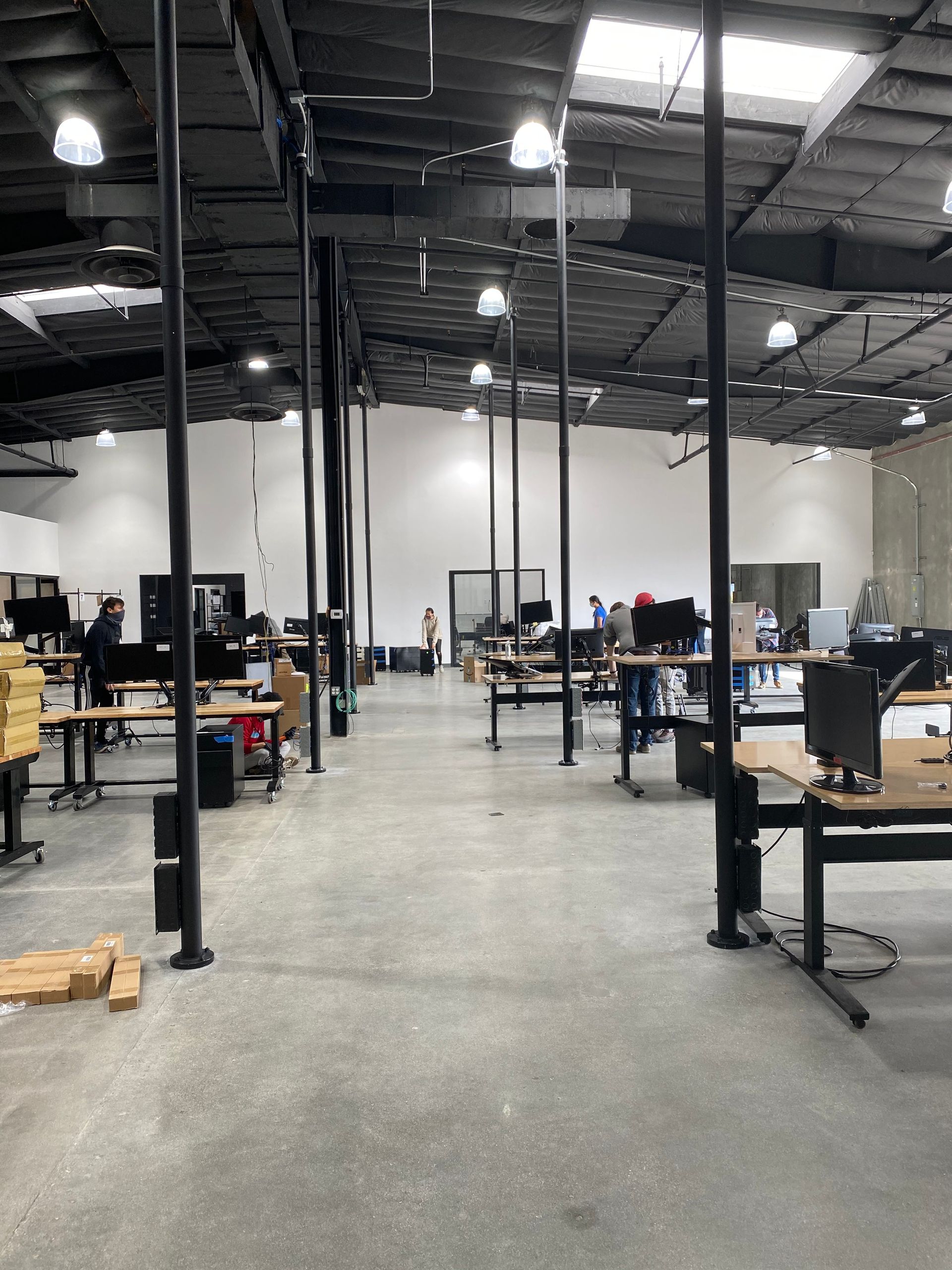 Large industrial workshop with desks, computers, tools, and tall black machinery under bright overhead lights.