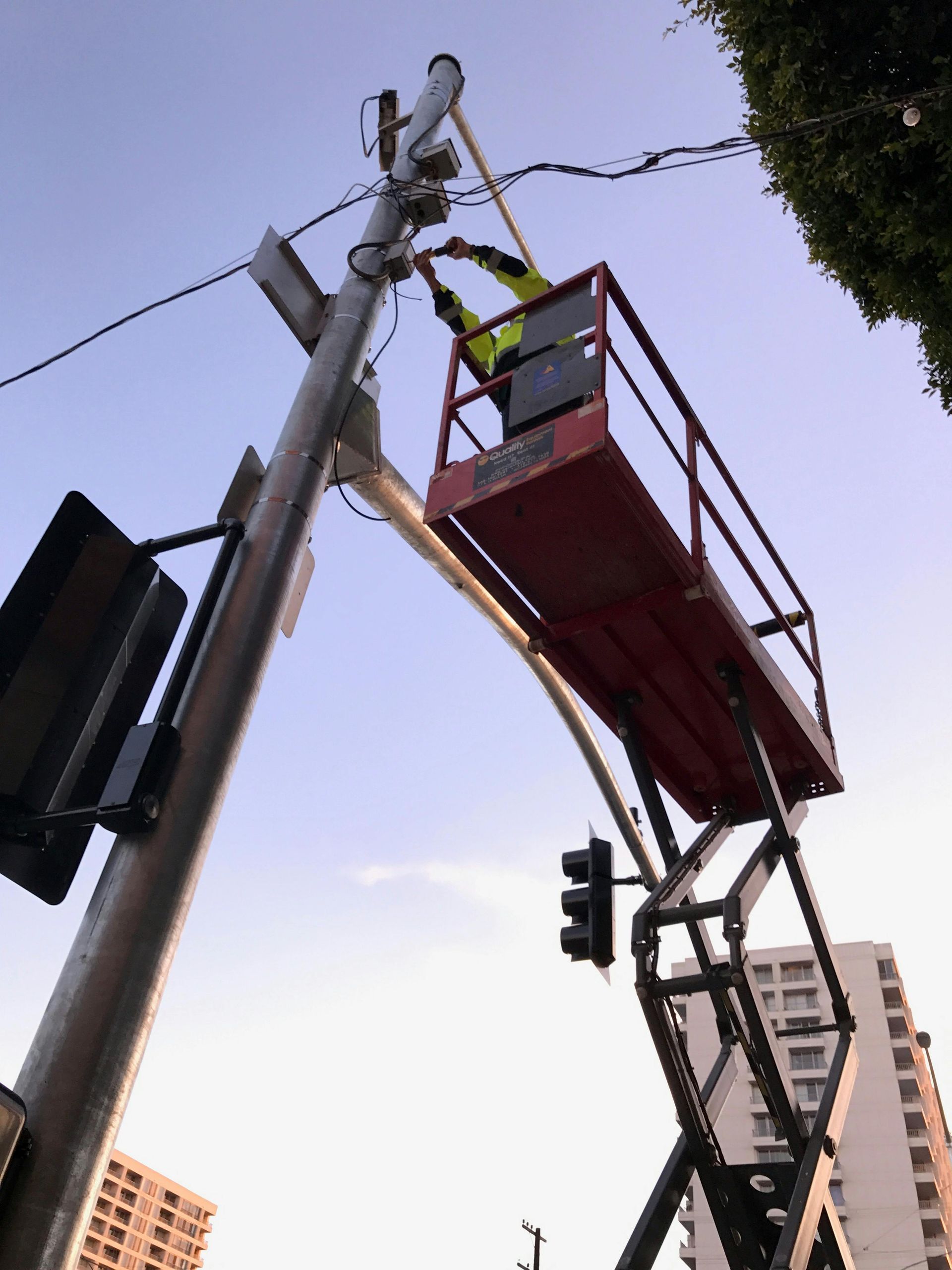 A worker in a yellow vest on a red scissor lift repairs wiring on a traffic light pole against a clear sky.