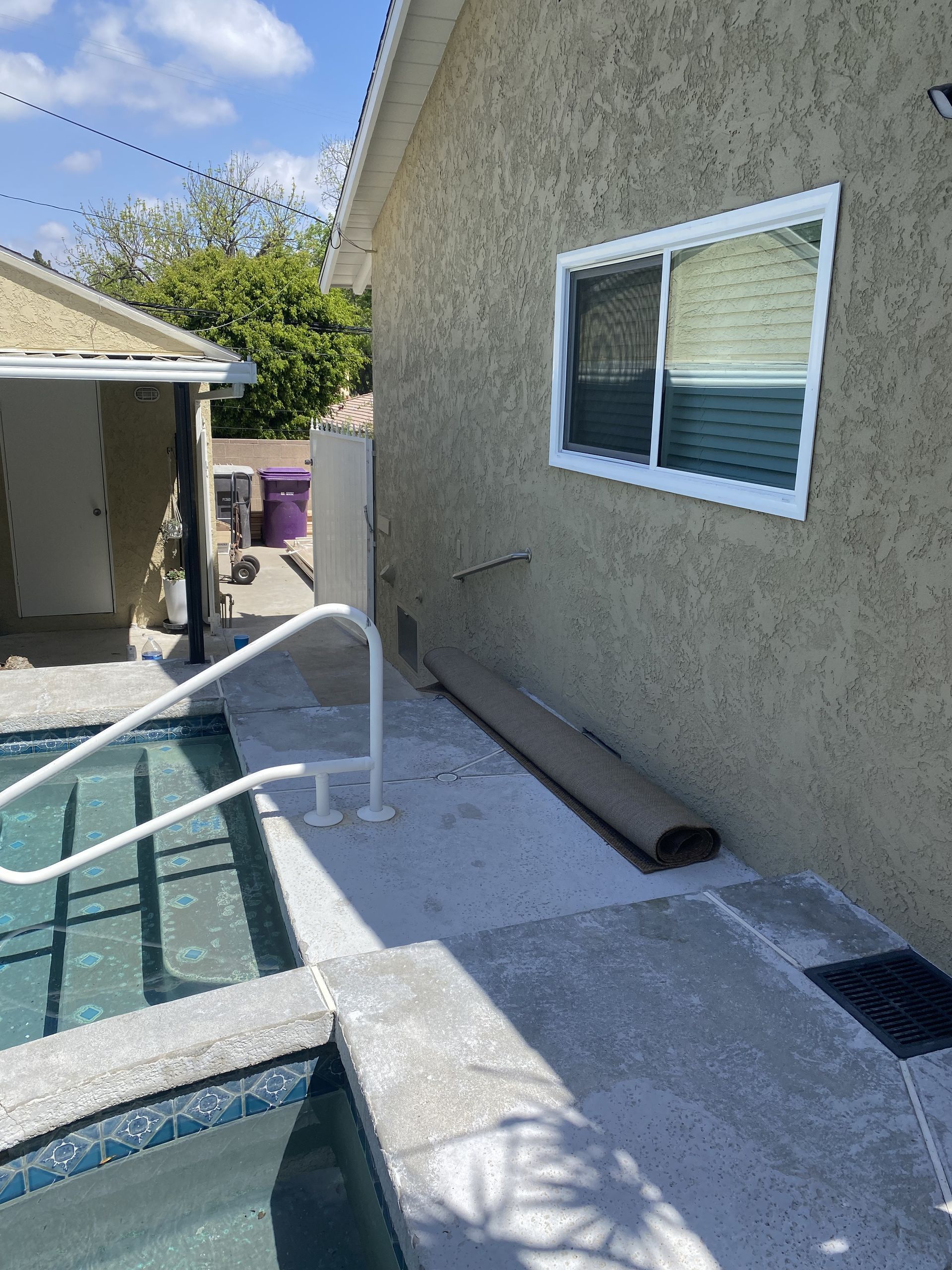 Backyard pool deck beside a stucco house wall with a window and drain grate