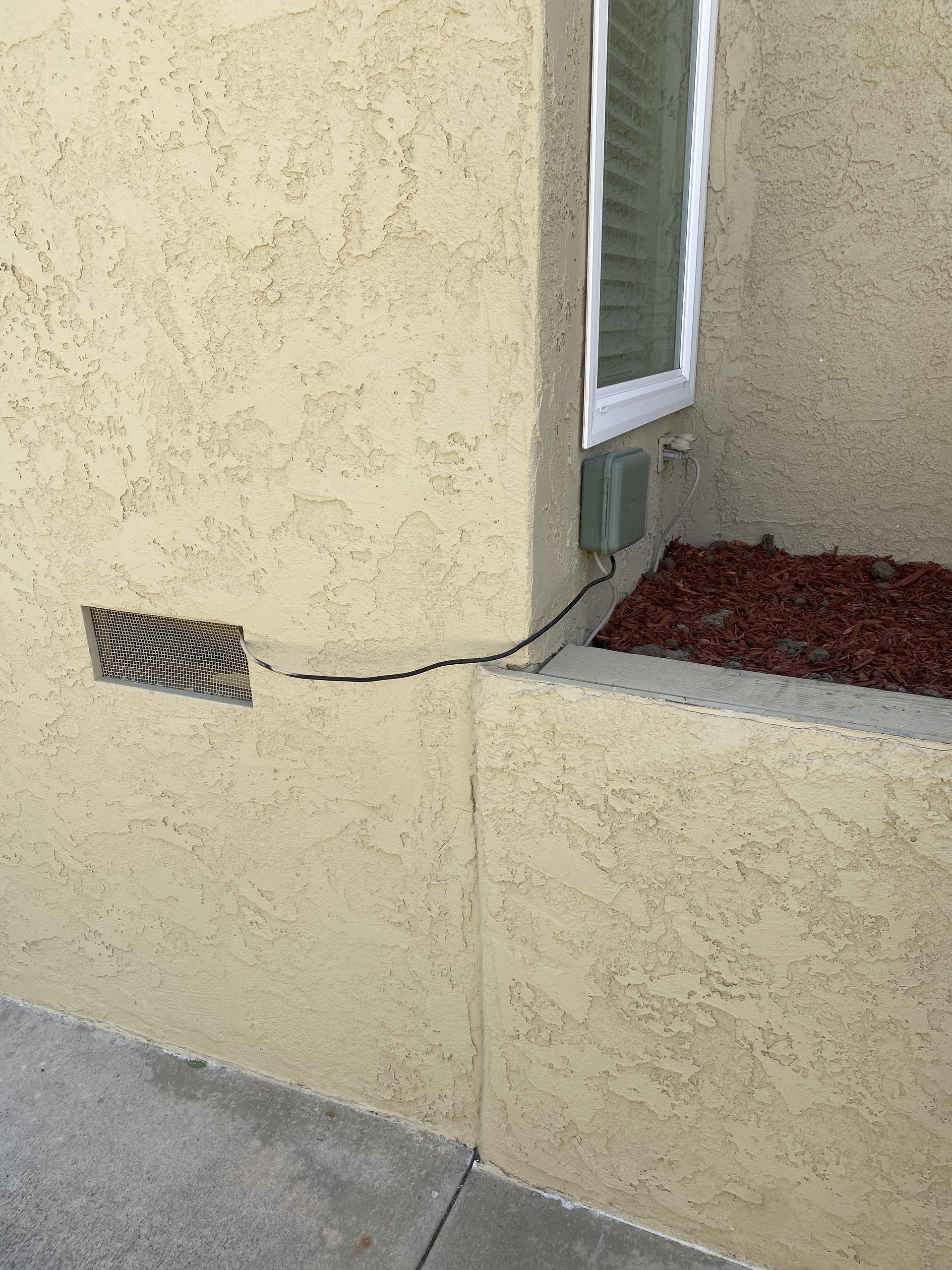 Exterior stucco wall with a vent, window, and a black cable running into a planter bed.