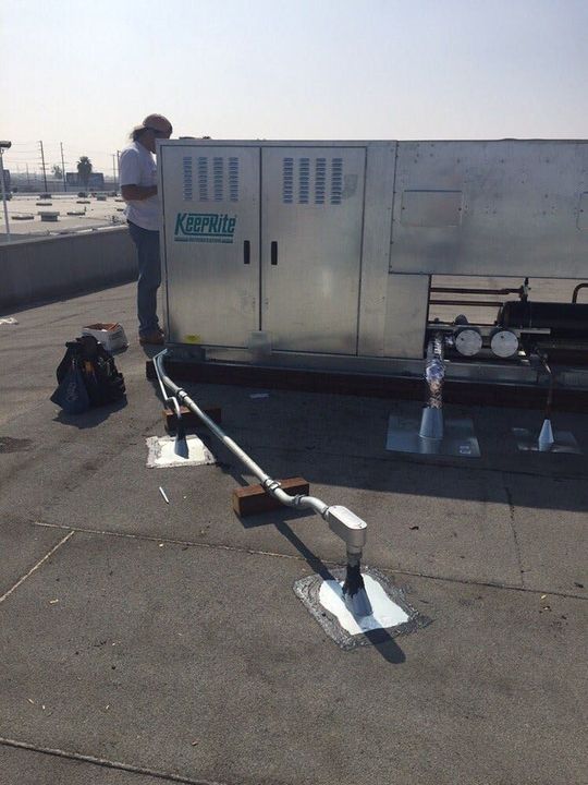 A technician stands next to an HVAC unit on a flat rooftop, inspecting a metal conduit connected to a sealed roof port.