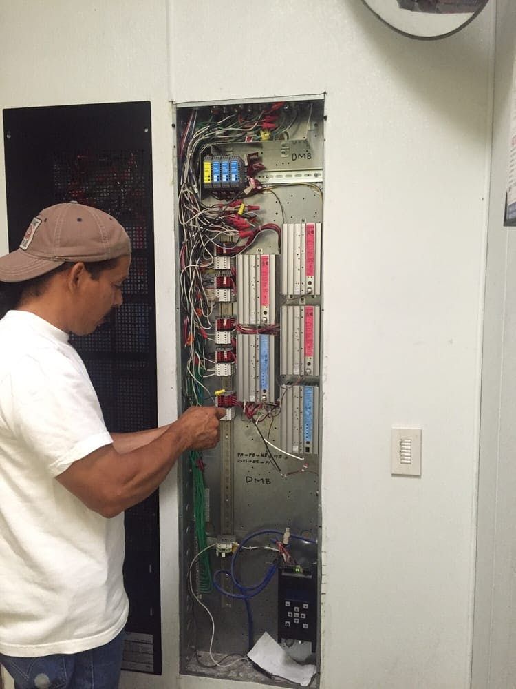 A person in a baseball cap works on an open wall-mounted electrical panel with several modules and wiring.