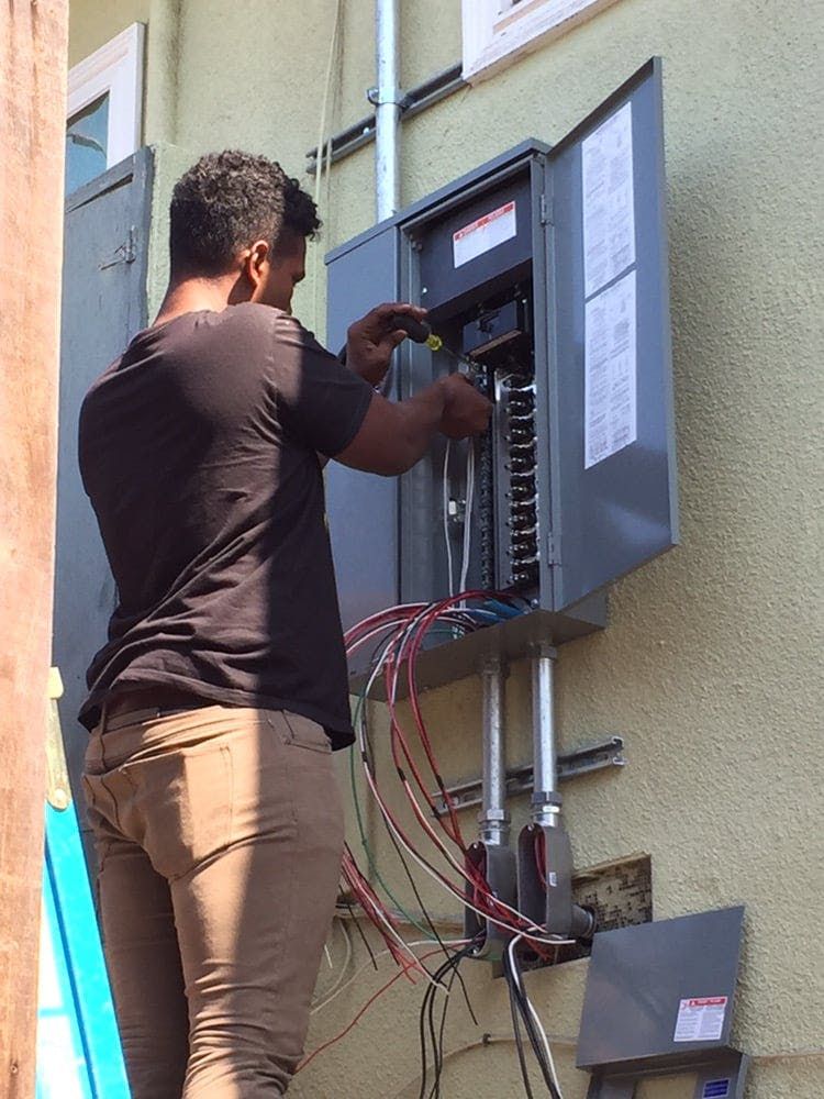 An electrician working on an open outdoor electrical panel, connecting wires to the breaker box on a stucco wall.