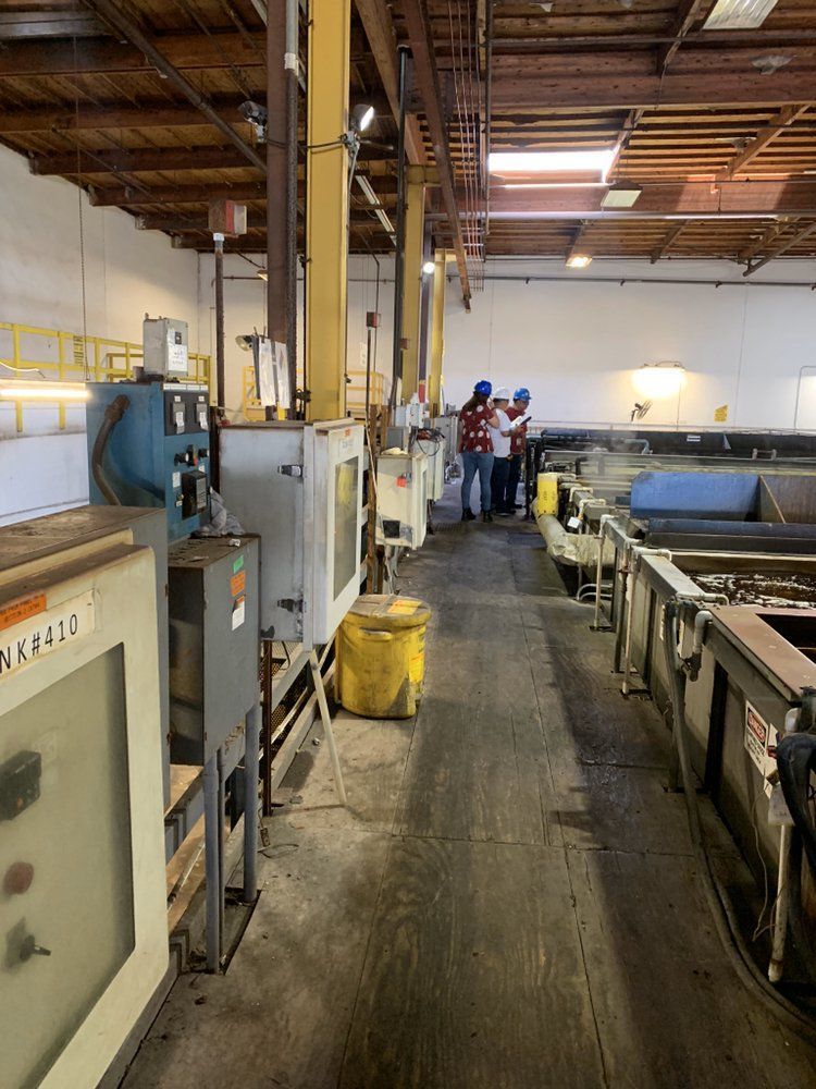 Three people in safety gear inspect machinery in a large industrial factory setting with a concrete aisle.