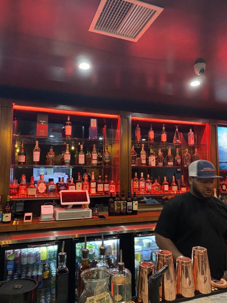 A bartender stands at a dimly lit bar with shelves filled with backlit liquor bottles and red ambient lighting.