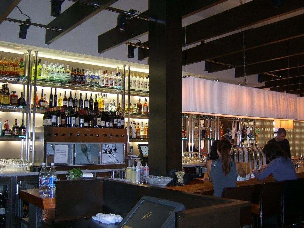 Patrons sit at a bar with a multi-tiered liquor display, illuminated by overhead pendant lights and exposed wood beams.