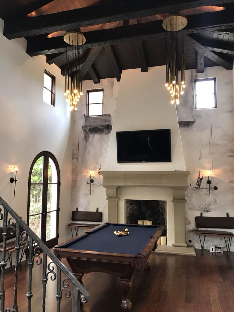 A pool table sits in a grand room with high ceilings, exposed wooden beams, large pendant lights, and a stone fireplace.